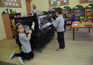 Dzieci malują na czarnej folii białą farbą zimowy krajobraz